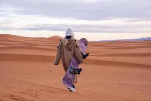 hombre vestido con ropa tradicional con sandboard caminando sobre dunas de arena contra el cielo foto