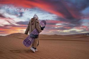 Man in traditional clothes with sandboard walking on sand dunes against sky photo