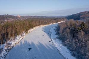 vista aérea del río y del bosque cubierto de nieve después de una ventisca en una neblina matutina. cielo azul claro. las maravillas de invierno. parque nacional de gauja, sigulda, letonia foto