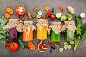 coloridos batidos y jugos saludables en botellas con frutas tropicales frescas y superalimentos sobre fondo de piedra oscura con espacio de copia. foto