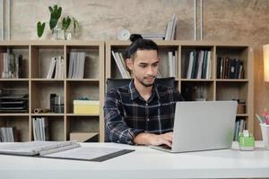Young male worker of Asian ethnicity uses laptop to do creative work on white desk in front of bookshelf of a casual workplace, startup business person, and online e-commerce occupation.