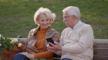 bella coppia senior seduta sulla panchina con cesto pieno di fiori e guardando il telefono cellulare video