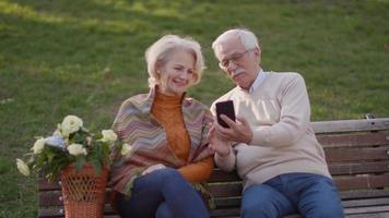 Handsome senior couple sitting on the bench with basket full of flowers and looking at mobile phone