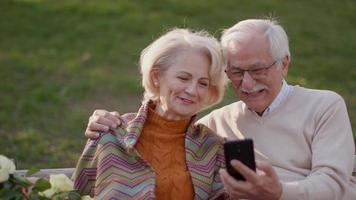 Handsome senior couple sitting on the bench with basket full of flowers and looking at mobile phone video