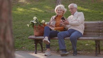 Handsome senior couple sitting on the bench with basket full of flowers and looking at mobile phone video