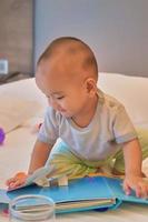 retrato de un feliz bebé asiático de 6 meses sentado en la cama y leyendo un libro foto