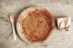 Dirty from food disposable paper utensils on a wooden background. Wooden fork and paper cup with plate on wooden background. Eco friendly disposable tableware. Top view. Copy, empty space for text photo