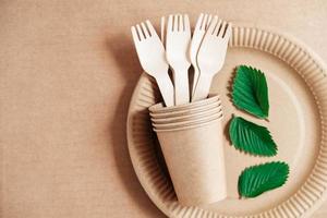 Wooden forks and paper cups with plates on kraft paper background. Eco friendly disposable tableware. Also used in fast food, restaurants, takeaways, picnics. Top view. Copy, empty space for text photo