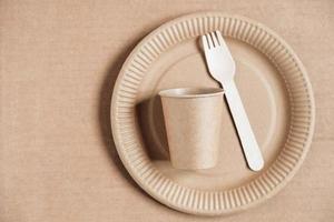 Wooden fork and paper cup with plate on kraft paper background. Eco friendly disposable tableware. Also used in fast food, restaurants, takeaways, picnics. Top view. Copy, empty space for text photo