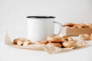 Metal mug with hot drink and mini round bagels on a white wooden background. Copy, empty space for text