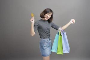 retrato de una joven y hermosa mujer asiática sosteniendo una tarjeta de crédito y una colorida bolsa de compras aislada sobre un estudio de fondo gris foto