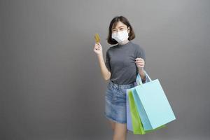 Portrait of young beautiful asian woman wearing a surgical mak is holding credit card and colorful shopping bag isolated over gray background studio