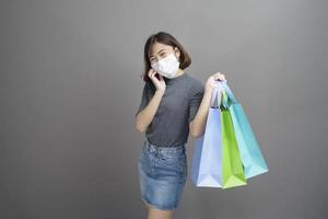 Portrait of young beautiful asian woman wearing a surgical mak is holding credit card and colorful shopping bag isolated over gray background studio