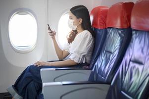 A young woman wearing face mask is using smartphone onboard, New normal travel after covid-19 pandemic concept photo