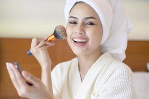 atractiva mujer feliz en albornoz blanco está aplicando maquillaje natural con cepillo de polvo cosmético, concepto de belleza.
