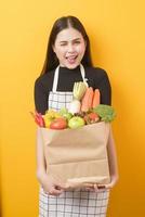 Beautiful young woman is holding vegetables in grocery bag in studio yellow  background photo