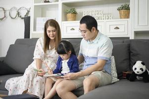 padre asiático feliz enseñando a su pequeña y linda hija a hacer la tarea y dibujar juntos en casa. foto