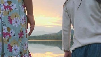 Close up of mother holding hands with her daughter on a summer day by the lake at sunset. Happy family spending time together on vacation. video