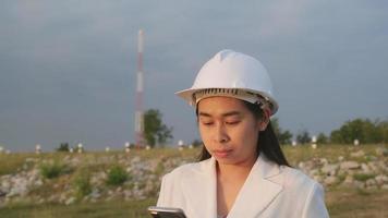 ingeniera que trabaja con un teléfono inteligente en el sitio de construcción de la represa para generar electricidad. mujer arquitecta segura de sí misma con casco blanco mirando el sitio de construcción de una presa. video