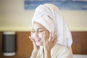 A happy beautiful woman in white bathrobe applying moisturizing cream on face in bedroom, skin care and treatment concept photo