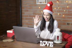 Young smiling woman wearing red Santa Claus hat making video call on social network with family and friends on Christmas day. photo