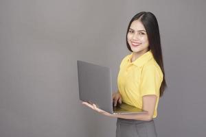 joven mujer hermosa y segura de sí misma con camisa amarilla sostiene una laptop en un estudio de fondo gris foto