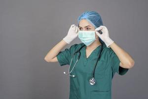 young confident woman doctor in green scrubs is wearing surgical mask over grey background studio photo