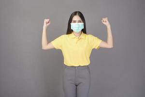 young confident woman in yellow shirt is wearing surgical mask over grey background studio photo