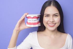 Young beautiful smiling woman is holding plastic denture teeth over isolated purple background studio photo