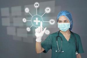 young confident woman doctor in green scrubs is wearing surgical mask over grey background studio , touching medical data on virtual screen , medical technology concept photo