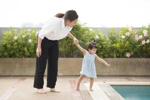 An Asian Mother is holding her baby girl hand to walking aside of pool , family, parenthood, home safety concept