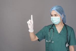 young confident woman doctor in green scrubs is wearing surgical mask over grey background studio photo