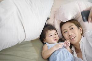 Asian Mother and her baby daughter are making selfie or video call to father in bed , Family , Home safety , parenthood , technology concept photo