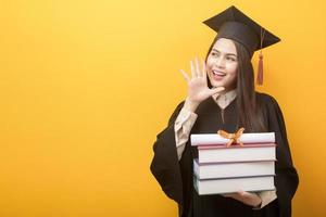 Beautiful woman in graduation gown is holding books and certificate on yellow background photo