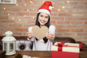 Young smiling woman wearing red Santa Claus hat showing a heart shaped model on Christmas day, holiday concept.