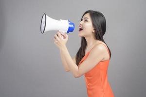 Fashion portrait of beautiful woman wearing orange dress is using megaphone isolated over gray background studio photo