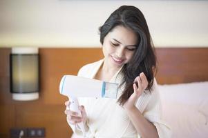 Smiling young woman wearing white bathrobe drying her hair with a hairdryer after a shower in bedroom photo