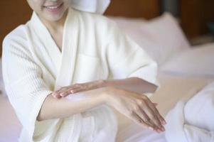 A happy beautiful woman in white bathrobe applying body moisturizing cream in bedroom, skin care and treatment concept photo