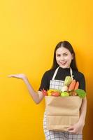 Beautiful young woman is holding vegetables in grocery bag in studio yellow  background photo