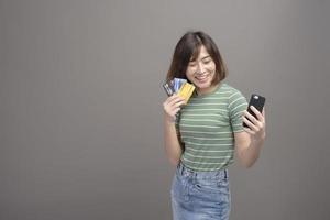 Portrait of young beautiful asian woman holding credit card and smartphone isolated over gray background studio photo