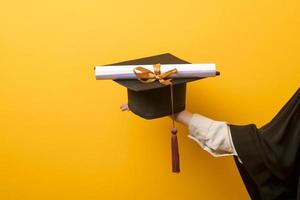 el primer plano de la mano de la mujer en la toga de graduación sostiene la gorra de graduación y el certificado sobre fondo amarillo foto