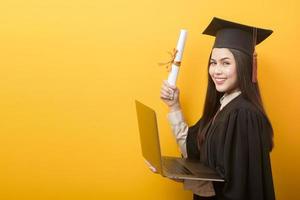 portrait of beautiful woman in graduation gown is holding laptop computer on yellow background photo