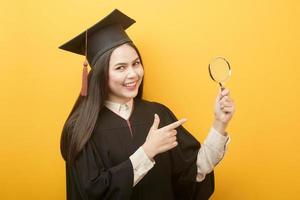 portrait of beautiful woman in graduation gown is holding magnifying glass on yellow background photo