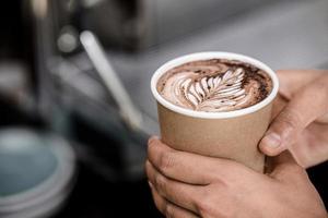 Close up shot of male hands holding take away cup of brewed hot coffee with Fern latte art design photo