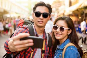 Smiling young Asian couple tourists taking selfie while traveling in Khao San road Bangkok Thailand during summer vacation