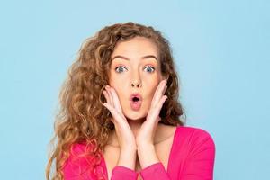 Surprised shocked caucasian woman with popped eyes and hands on cheeks in blue background photo