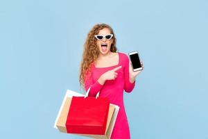Shocked woman with colorful shopping bags yelling and pointing to mobile phone isolated on blue background