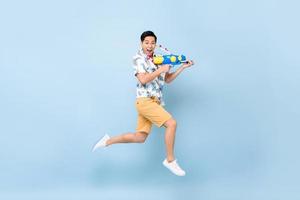 apuesto joven asiático sonriente jugando con pistola de agua y saltando en el fondo azul del estudio para el festival songkran en tailandia y el sudeste asiático foto