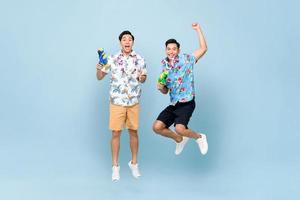 Smilng happy Asian male friends playing with water guns and jumping in blue isolated background for Songkran festival in Thailand and southeast Asia photo