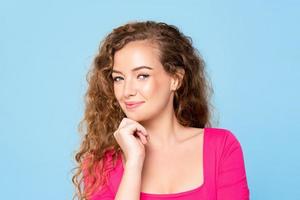 Waist up portrait of pretty happy caucasian woman in pink blouse smiling with hand on chin isolated on light blue background